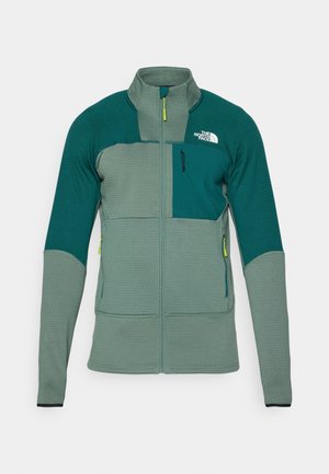 Grüne Outdoor-Jacke aus strukturiertem Stoff, hoher Kragen, Frontreißverschluss und kontrastierende Farbpaneele mit einer Brusttasche und Logo.
