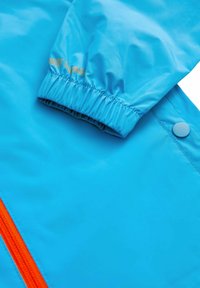 Jasnoniebieska wodoodporna kurtka z elastycznym mankietem, pomarańczowym zamkiem i złotym napisem na rękawie dla dodatkowego efektu.