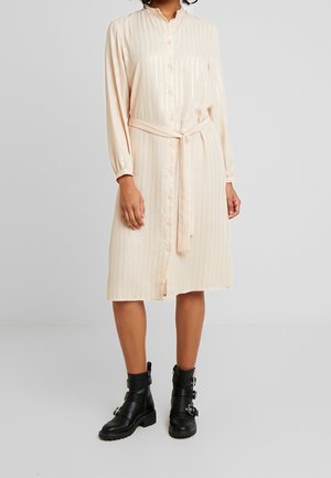Mujer con un vestido rosa pálido hasta la rodilla, abotonado, con mangas largas y cinturón, combinado con botas negras hasta media pantorrilla con hebillas.