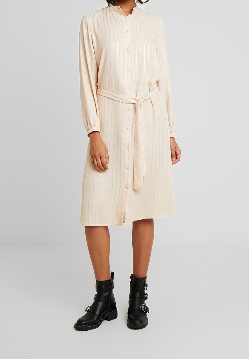 Femme portant une robe rose pâle boutonnée jusqu’au genou, manches longues et ceinture, associée à des bottes noires à mi-mollet avec boucles.