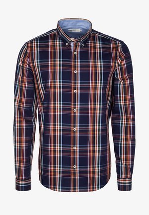 Man draagt een opgerolde geruite blouse, zwarte spijkerbroek en zwarte laarzen, houdt een opgevouwen zwart gewatteerd jack in zijn rechterarm.