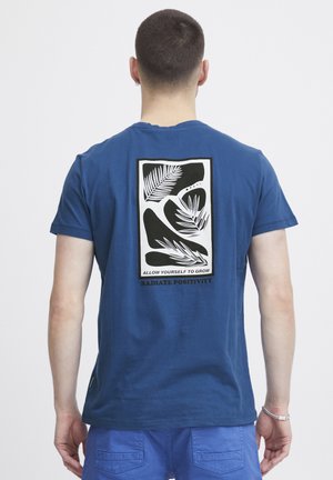 Blue cotton t-shirt with a black and white graphic design featuring abstract leaf patterns and text on the back. Short sleeves, crew neck.