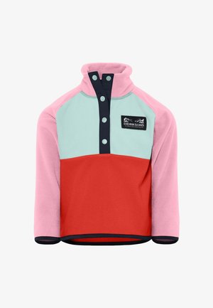 Enfant aux cheveux blonds bouclés portant une veste en polaire colorée avec des panneaux rose, rouge et bleu clair, un pantalon en polaire rouge assorti et des chaussettes blanches.