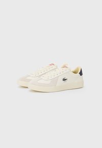Cream-colored sneakers with a suede toe cap, lace-up design, and small black logo on the side. Features red accents on the tongue.