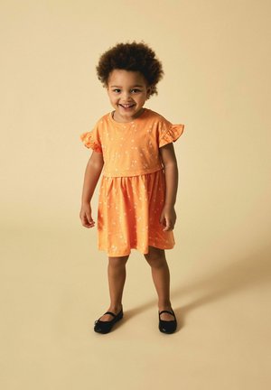 Vestido corto de manga corta naranja con patrones florales y mangas acampanadas. Bailarinas negras, de pie sobre un fondo beige claro.