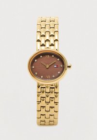 Złototonowana bransoleta zegarka połączonego z owalną brązową tarczą, kryształowymi znacznikami godzin, cyframi rzymskimi na 12 i 6 oraz małym logo w kolorach czerwonym, białym i niebieskim.