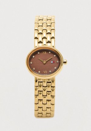 Złototonowana bransoleta zegarka połączonego z owalną brązową tarczą, kryształowymi znacznikami godzin, cyframi rzymskimi na 12 i 6 oraz małym logo w kolorach czerwonym, białym i niebieskim.