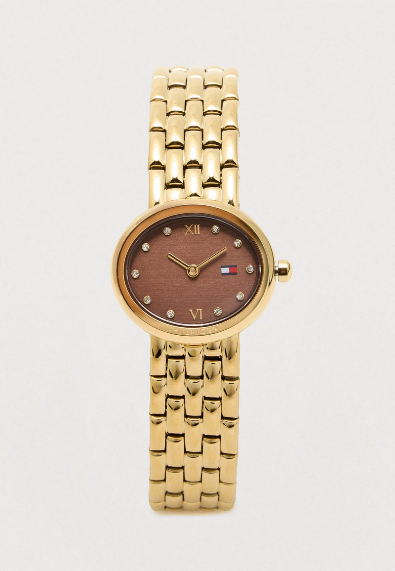 Złototonowana bransoleta zegarka połączonego z owalną brązową tarczą, kryształowymi znacznikami godzin, cyframi rzymskimi na 12 i 6 oraz małym logo w kolorach czerwonym, białym i niebieskim.