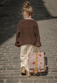 Ljusrosa ryggsäck med röda fjärils- och jordgubbsmönster, gula remmar, bärs av ett barn som har en brun stickad tröja och vita byxor.