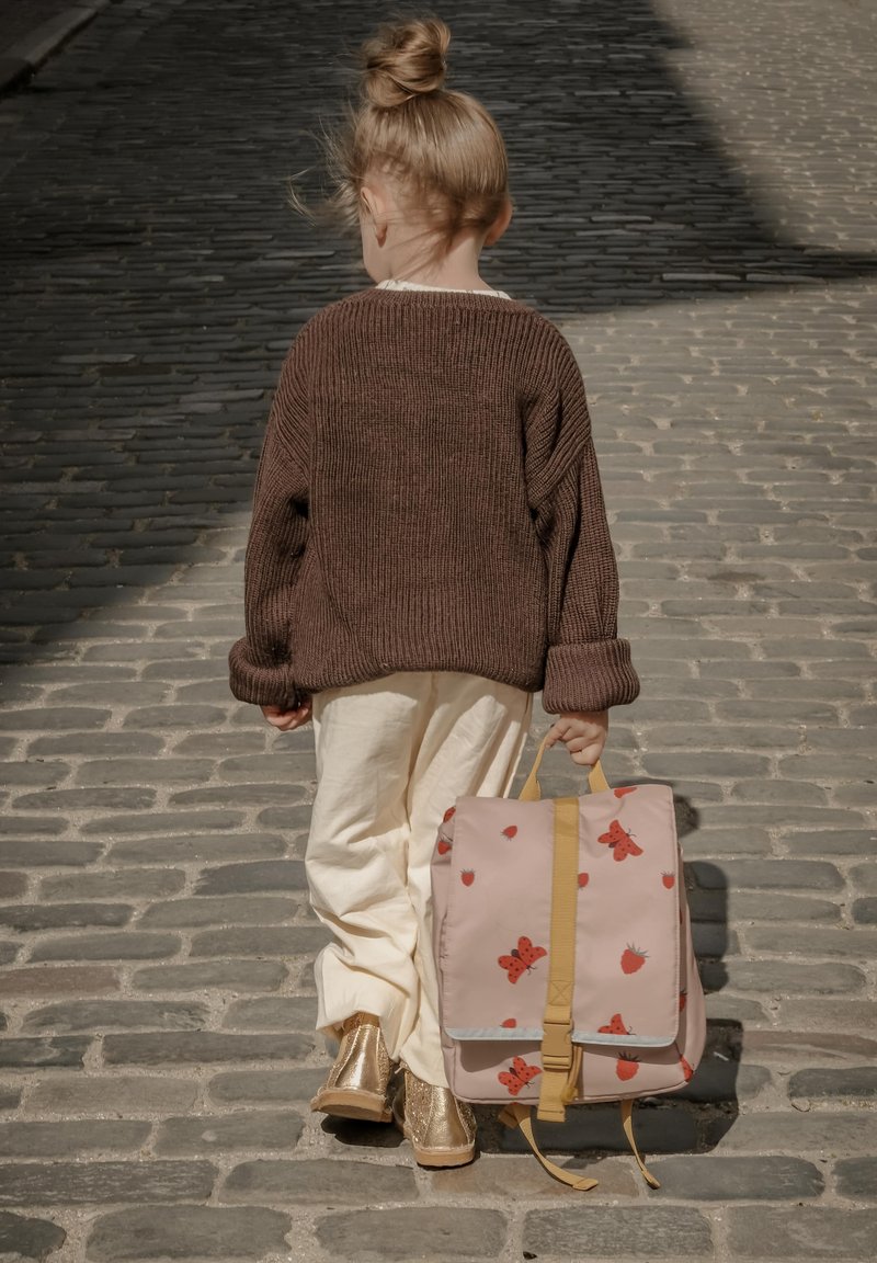 Ljusrosa ryggsäck med röda fjärils- och jordgubbsmönster, gula remmar, bärs av ett barn som har en brun stickad tröja och vita byxor.