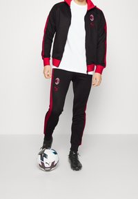 Schwarzer Trainingsanzug mit roten Akzenten, bestehend aus einer Reißverschlussjacke und passenden Hosen. Mit Logodetails. Person, die einen Fußball hält.