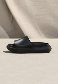 Μαύρο λαστιχένιο σανδάλι τύπου σαγιονάρας με χοντρή σόλα και ανοιχτά δάχτυλα, τοποθετημένο σε φωτεινή επιφάνεια με φόντο από μαλακό ύφασμα.