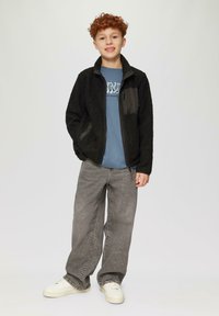 Zwarte fleecejack met een ritsborstzak, gedragen over een blauw T-shirt, gecombineerd met grijs wijde jeans en witte sneakers.