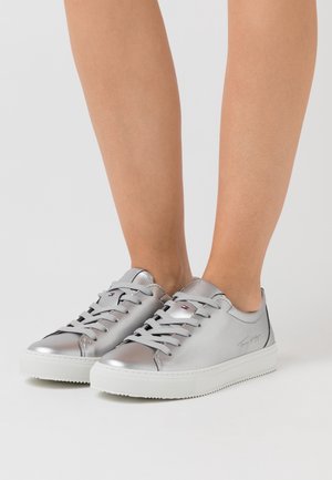 Silberne Sneaker mit niedrigem Schnitt und weißen Sohlen, getragen an nackten Füßen, mit grauen Schnürsenkeln und dezentem Markenlogo an der Seite und der Zunge.