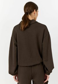 Hnědý, oversized pulovr s měkkou texturou, spadlými rameny a žebrovanými manžetami. Záda mají jednoduchý, čistý design bez ozdob.