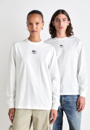 Balti ilgomis rankovėmis marškiniai, pagaminti iš medvilnės, su mažuoju Lacoste logotipu ant krūtinės. Vienas modelis dėvi smėlio spalvos kelnes, kitas - mėlynus džinsus.