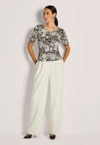 Vrouw in een zwart-witte bloemen top met korte mouwen, wijde crèmekleurige broek en donkere puntschoenen, staand met handen in de zakken.