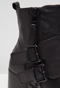 Gros plan sur une bottine noire en cuir avec une fermeture éclair latérale et deux brides réglables à boucle sur un talon superposé.