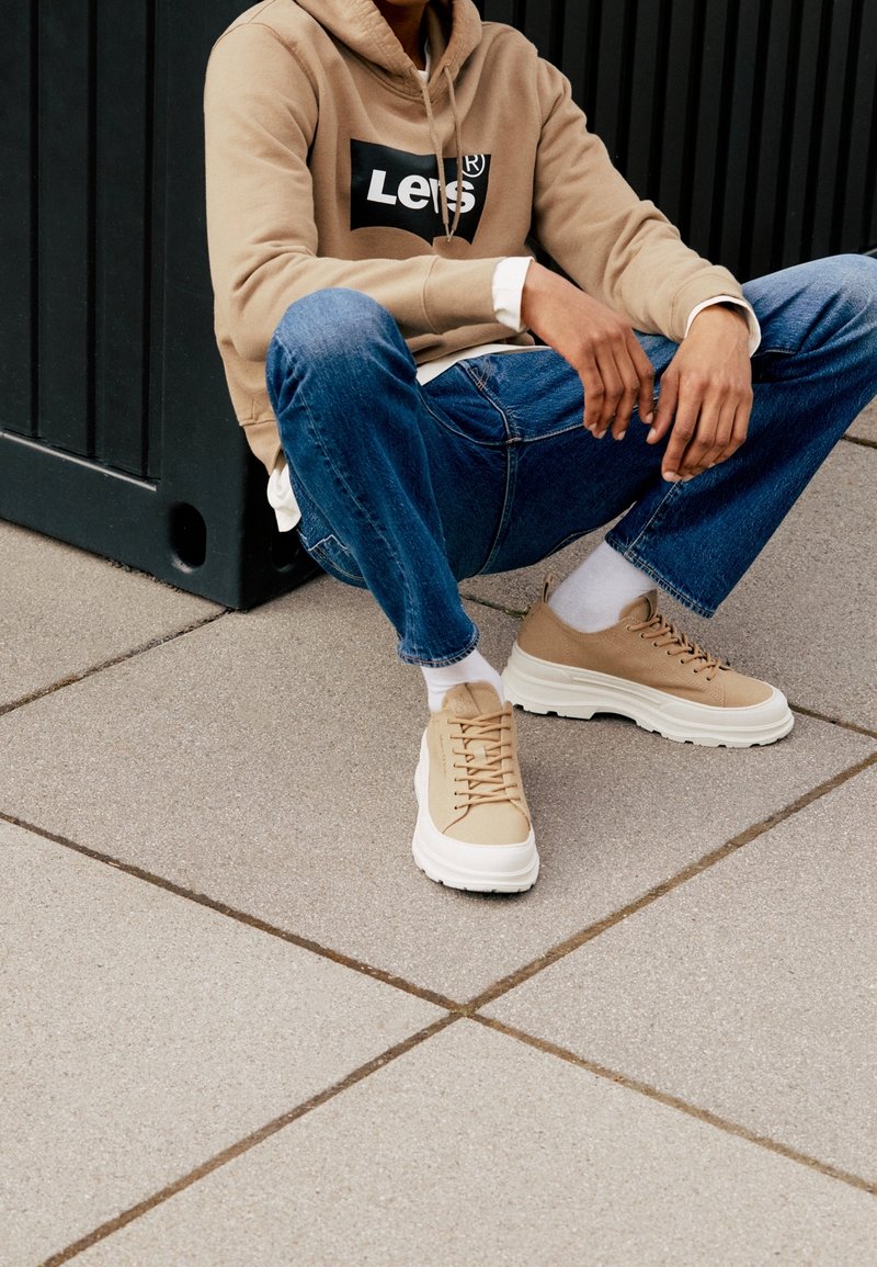 Personne portant un sweat à capuche beige Levi's, un jean bleu, des chaussettes blanches et des baskets beiges assise sur le trottoir contre un mur noir.