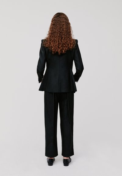 Blazer nero su misura con spalle strutturate abbinato a pantaloni a gamba larga coordinati e scarpe nere lucide, visto da dietro.