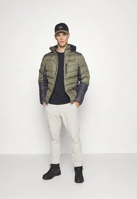 Alyvuogių žalios spalvos pūkinė striukė su tamsiai mėlynomis rankovėmis, užtrauktuku užsegama kišene, juoda marškinėliai, šviesiai smėlio spalvos kelnės ir juodi batai. Modelis stovi užtikrintai.