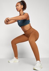 Nike sportski grudnjak u tirkiznoj boji, visoki smeđi tajice i bijele sportske cipele. Odjeća ima uski kroj i tkaninu koja upija vlagu.
