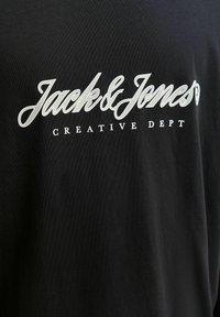 Μαύρο ύφασμα με λευκό κείμενο που γράφει "Jack & Jones" με μεγάλη καλλιγραφική γραμματοσειρά και "CREATIVE DEPT" με μικρότερα κεφαλαία γράμματα από κάτω.