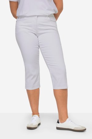 Persoon die witte mid-kuitbroeken draagt, witte sneakers met zwarte accenten en een wit overhemd,staand met de handen achter de rug.