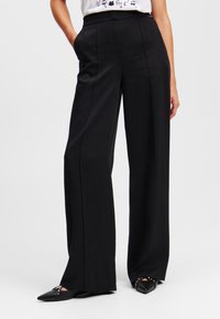 Zwarte wijde broek gemaakt van een gladde stof, met een taillering ontwerp, voorplooien en zijzakken. Gemaakt met puntige zwarte flats.