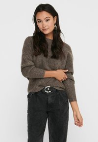 Pull en maille marron à coupe ample et avec des poignets côtelés, associé à un jean noir taille haute et une boucle de ceinture circulaire. Boucles d'oreilles simples visibles.