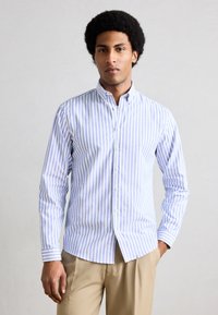 Άνδρας με μαύρα, σγουρά μαλλιά, φοράει μπλε και λευκό πουκάμισο με κάθετες ρίγες και μπεζ παντελόνι, στέκεται μπροστά σε ουδέτερο φόντο.