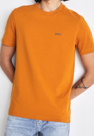 Man draagt een feloranje T-shirt met korte mouwen en het "BOSS"-logo op de borst, en lichte blauwe jeans, met een stierschedel-tatoeage op de rechteronderarm.