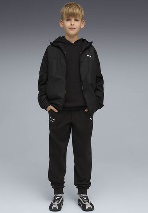 Ragazzo in piedi con le mani in tasca che indossa una giacca nera Puma, pantaloni neri con il logo BMW Motorsport e sneakers Puma nere e bianche.