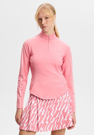 Donna bionda con coda di cavallo che indossa una maglia rosa a maniche lunghe con cerniera e una gonna plissettata rosa e bianca con motivo a fantasia.