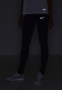 Osoba stoi w ciemnych legginsach, jasnych sneakersach i jasnej bluzce, wszystkie z widocznymi odblaskowymi logo Nike w słabym świetle.