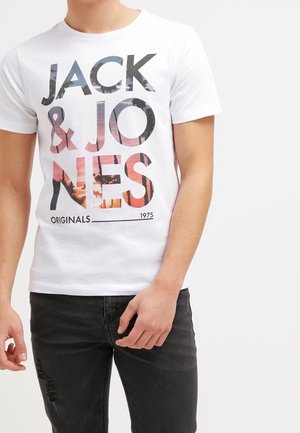 Mężczyzna ubrany w białą koszulkę z dużym napisem "JACK & JONES ORIGINALS 1975" w kolorach inspirowanych zachodem słońca oraz czarne podniszczone dżinsy.