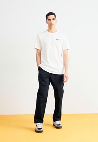 Camiseta de algodón blanca con cuello redondo y pequeño logo, combinada con pantalones negros. El modelo lleva zapatillas en blanco y negro.