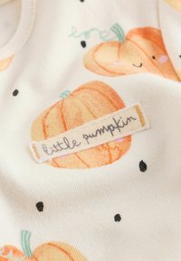 White fabric with smiling orange pumpkin illustrations and a sewn label reading "little pumpkin" in cursive handwriting.