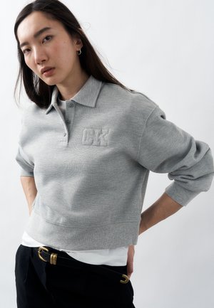 Mladá žena s dlhými tmavými vlasmi nosí sivú mikinu s golierom a logom "CK", biele tričko pod ňou, čierne nohavice a opasok so zlatou prackou.