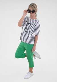 Gestreepte zwart-witte t-shirt met grafische tekst, gecombineerd met groene cropped broek en witte sportschoenen. Zonnebril maakt de look compleet.