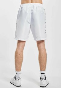 Hvit treningsshorts med elastisk liv, med svarte sideaksenter og et glatt, lett stoff. Kombinert med hvite joggesko.