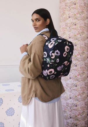 Rugzak met bloemmotief in marineblauw met roze en lavendelbloemen, met een afgeronde vorm en een bovenste draaglus, gedragen over een beige jas.