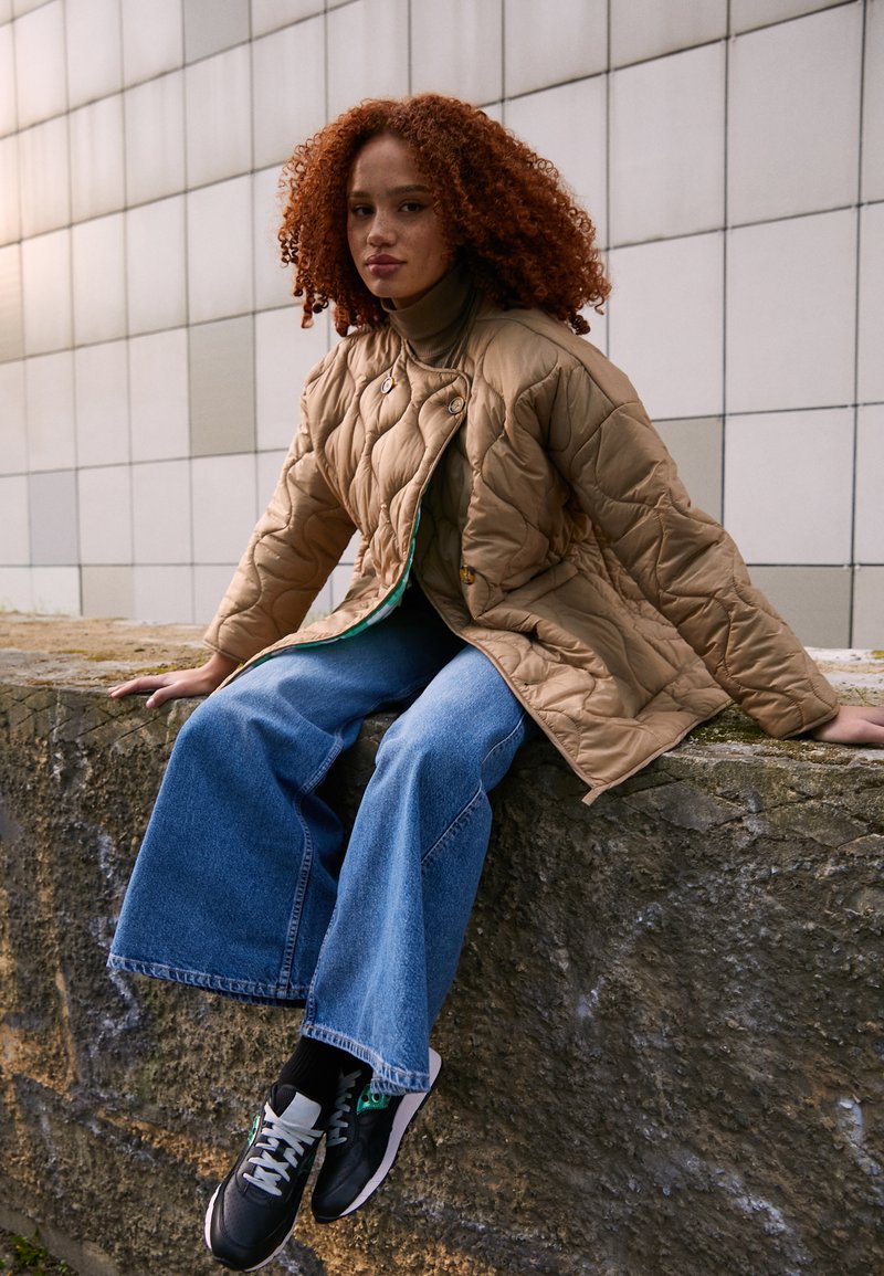 Beige gesteppte Jacke mit runden Knöpfen, grünem Futter; olivgrüner Rollkragenpullover, weite blaue Jeans, schwarz-weiße Sneakers, sitzend auf einer Steinmauer.