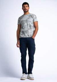 Šedé tričko s texturovaným vzorem, oblečené s námořnickými cargo kalhotami. Model stojí ve sneakers, ruce v kapsách, na jednobarevném pozadí.