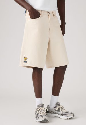 Hombre con pantalones cortos beige hasta la rodilla con logo de gallo en la pierna izquierda, calcetines blancos, zapatillas Nike grises y blancas y camisa blanca.