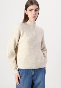 Beige, ribbestrikket genser med høy krage og glidelås, med lange ermer og en avslappet passform; kombinert med blå denimjeans.
