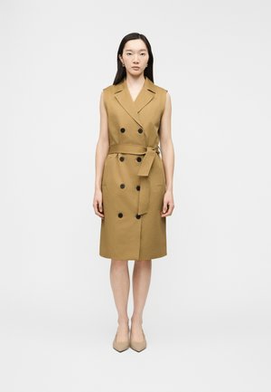 Donna che indossa un vestito trench senape senza maniche con bottoni neri e cintura in vita, in piedi davanti a uno sfondo bianco semplice.