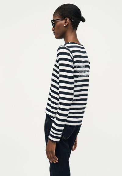 Γυναίκα με σκούρο δέρμα και μαλλιά σε κότσο φοράει μαύρα γυαλιά ηλίου, μια μαύρο-λευκή ριγέ μπλούζα με μακριά μανίκια και σκούρα τζιν, στεκόμενη στο πλάι.