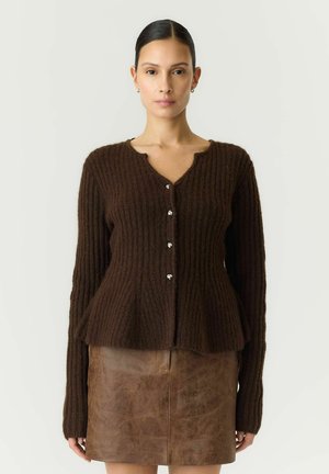 Donna con capelli scuri in cardigan marrone a coste con bottoni e gonna in pelle marrone, in piedi davanti a uno sfondo chiaro uniforme.