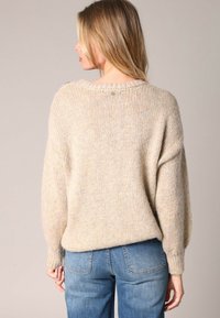 Pull en maille beige au fit ample, avec des épaules tombantes et un col en côtes. Texture douce avec des variations de couleur subtiles. Associé à un jean bleu.